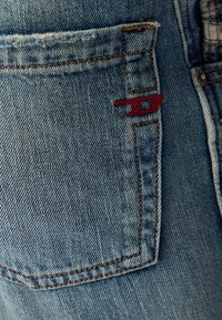 Pantalones vaqueros de denim en un color azul desgastado, con un bolsillo trasero que presenta un logo bordado en rojo y detalles de costura en contraste.