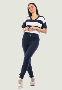 Pantaloni aderenti blu navy con una texture liscia, abbinati a una maglietta bianca a maniche corte a righe con scollatura a V. Le sneakers sono beige.