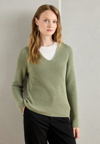 Oliwkowy sweter z ribbingu z dekoltem w kształcie V, długimi rękawami i teksturowanym wykończeniem, połączony z białą bluzką i czarnymi spodniami.