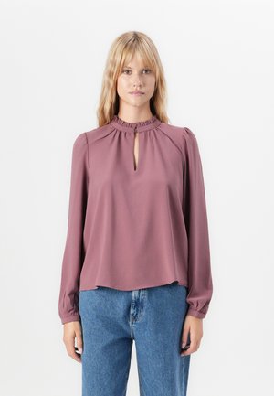 Junge Frau, die eine blassrosafarbene Bluse mit langen Ärmeln und einem geknöpften Ausschnitt sowie eine blaue Jeans trägt, vor einem weißen Hintergrund steht.