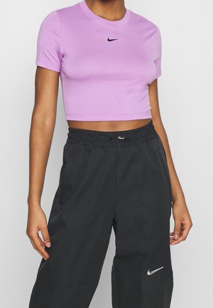 Osoba ubrana w liliowy krótki T-shirt Nike i luźne czarne spodnie Nike z widocznymi logo, stojąca na jednolitym jasnym tle.