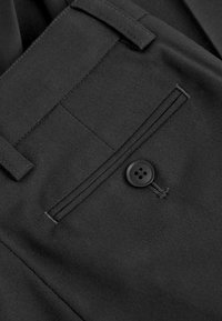 Pantalon noir sur mesure avec une texture lisse, doté d'une poche latérale avec une fermeture à bouton noir et des détails de couture subtils.