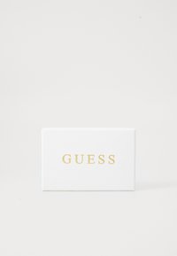 Biely obdĺžnikový box s textúrovaným povrchom, s nápisom "GUESS" v výraznom zlatom písme na prednej strane. Jednoduchý a minimalistický dizajn.