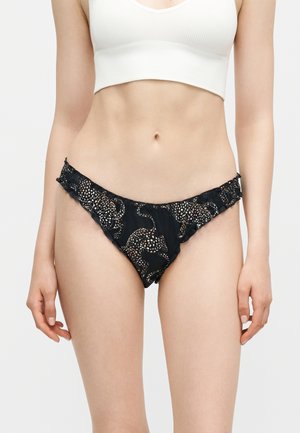 Kvinde iført sort bikinibund med leopardmønster og hvid cropped ribbet tanktop mod en ensfarvet baggrund.