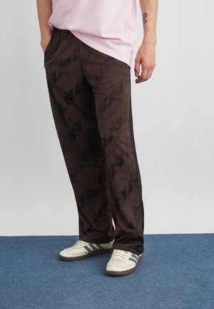 Persona che indossa pantaloni della tuta marrone scuro a fantasia con righe laterali, sneakers bianche con strisce nere e una camicia rosa chiaro.