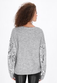 Szary sweter dzianinowy z długimi rękawami, ozdobiony srebrnymi cekinami, o miękkiej fakturze i luźnym kroju. Widok z tyłu, ukazujący projekt rękawów.