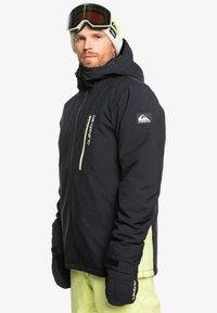 Schwarze wasserdichte Jacke mit einem gelben Reißverschluss, thermischen Handschuhen und einem Emblem am Ärmel; kombiniert mit hellgelben Hosen und Skibrille.
