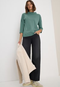 Camiseta de manga larga color teal con cuello alto, combinada con pantalones negros holgados y una chaqueta de forro polar color crema. Los accesorios incluyen zapatillas de deporte de color claro.