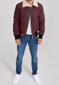 Mężczyzna w bordowej kurtce z kołnierzem z owczej wełny, białej koszulce, niebieskich dżinsach i białych trampkach stojący na jasnym tle.