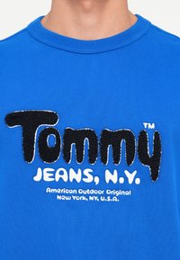 Felpa in cotone blu con un logo ricamato nero testurizzato, "Tommy," e la scritta "JEANS, N.Y." in bianco sottostante.
