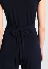 Combinaison bleu marine à manches courtes, taille cintrée et ceinture en tissu nouée au dos. Matière lisse et extensible sans motifs apparents.