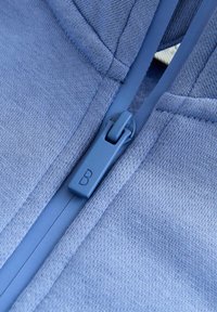 Prim-plan cu un fermoar albastru pe o țesătură texturată, prezentând cârligul fermoarului cu litera "B" și detalii ale cusăturii țesăturii.