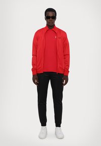 Sarkans zip-up džemperis virs sarkanās T-kreklu, apvienots ar melniem biksēm un baltām kedām. Modelis valkā saulesbrilles un ir ar īsiem matiem.