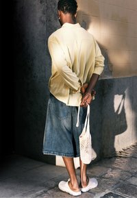 Camisa de manga larga amarilla clara, pantalones cortos de mezclilla azules, zapatos blancos tipo slip-on y una bolsa de red beige. Persona de pie con las manos detrás de la espalda.
