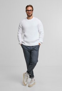 Wit kabelgebreid sweater gecombineerd met grijze op maat gemaakte broek en sneakers met beige accenten. Model staat met de armen gekruist. Eenvoudige achtergrond.