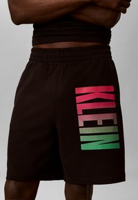 Short noir avec une taille élastique portant le nom de la marque "KLEIN" en grandes lettres dégradées du rouge au vert sur la cuisse gauche.