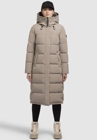 Beige pufferjas met een lange snit, voorzien van een capuchon, een rits aan de voorkant en twee zijzakken. Gedragen over zwarte legging en crèmekleurige laarzen.