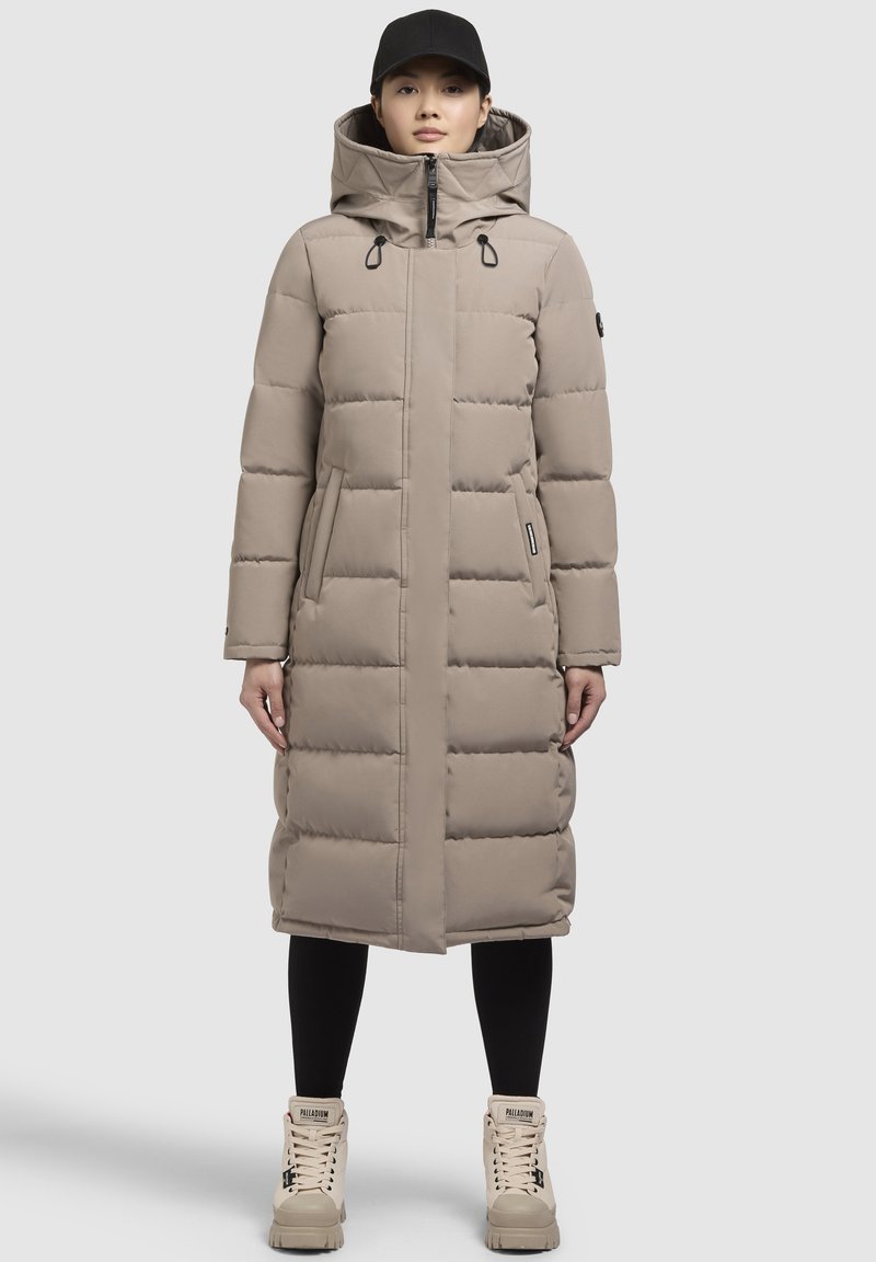 Beige pufferjas met een lange snit, voorzien van een capuchon, een rits aan de voorkant en twee zijzakken. Gedragen over zwarte legging en crèmekleurige laarzen.