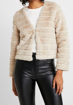 Kobieta ubrana w beżową, pasiastą kurtkę z futerka sztucznego na białej koszuli, w połączeniu z czarnymi, skórzanymi spodniami z wysokim stanem, stojąca na białym tle.