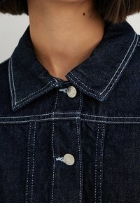 Tummansininen farkkutakki, jossa on kaulus, hopeanväriset napit, erottuva valkoinen tikkaus ja teksturoitu pinta. Lähikuva yläosasta.