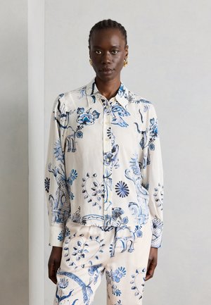 Frau trägt weißes Hemd mit Knöpfen und passende Hose mit blauem Blumen- und abstraktem Muster, steht vor einer schlichten hellen Wand.