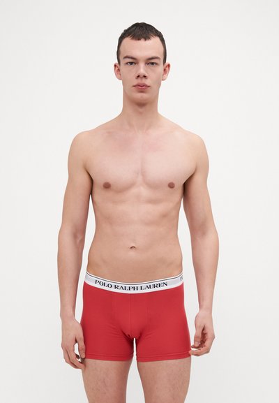 Crvene bokserice od mekog pamuka, s bijelim elastičnim pojasom na kojem je otisnuto "POLO RALPH LAUREN" u crnoj boji. Glatka tekstura, prilagođen dizajn.