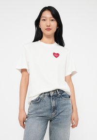 Mujer joven vistiendo una camiseta blanca con un logo de corazón rojo y jeans azul desgastado, de pie contra un fondo blanco liso.