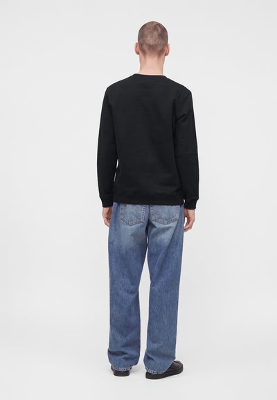 Homme debout de dos, portant un sweat-shirt noir, un jean bleu ample et des chaussures noires sur un fond clair uni.
