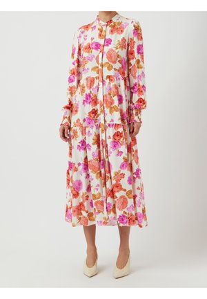 Femme portant une robe midi blanche à manches longues avec un imprimé floral rose, orange et marron, associée à des talons pointus crème.