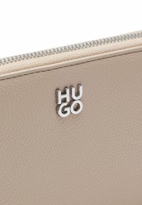 Beżowy skórzany portfel z zamkiem błyskawicznym, fakturowaną powierzchnią oraz srebrnym logo "HUGO" na froncie.