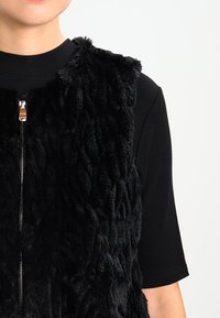 Svart konstpälsväst med en strukturerad yta, med en frontdragkedja och hög halsringning, i kombination med en svart ribbad topp under.