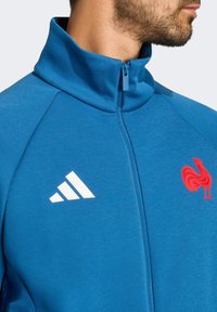 Modrá športová bunda s vysokým golierom, zipsom, bielym logom Adidas a červeným emblémom kohúta; má hladký, elastický materiál.