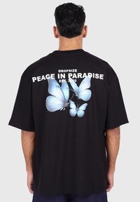 Black oversized t-shirt featuring large blue butterfly graphics on the back, with white text reading "DROPSIZE PEACE IN PARADISE EST. 2020."