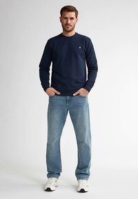 Bluză bleumarin cu guler rotund, din bumbac, purtată cu blugi bleu deschis. Are un logo mic pe piept și manșete striate. Adidași albi.