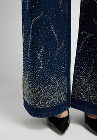 Pantalones vaqueros de pierna ancha en azul marino oscuro, con adornos de pedrería plateada en patrones ondulados en los muslos y el dobladillo.
