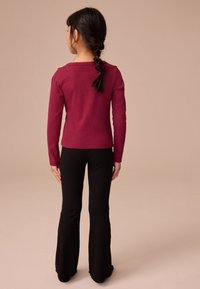 Majica z dolgimi rukavi v bordo rdeči barvi z ohlapnim krojem, kombinirana s črnimi rebrastimi hlačami z zvončastimi hlačnicami, ki imajo teksturirano tkanino in razširjen rob.