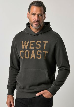 Mężczyzna z krótkimi włosami i brodą nosi czarną bluzę z kapturem z napisem "WEST COAST", stoi z jedną ręką w kieszeni na jednolitym tle.