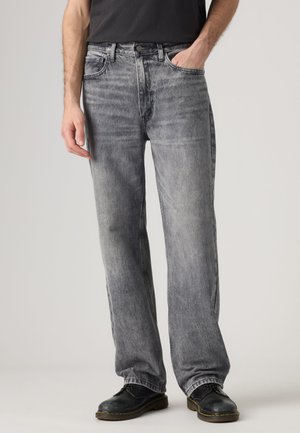 Mann trägt ausgewaschene graue Jeans mit geradem Bein, schwarze Schuhe und ein schwarzes T-Shirt, mit einer Hand in der Tasche vor einfachem Hintergrund.