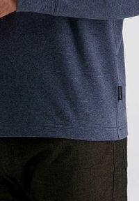 Nahaufnahme einer Person, die ein blaues strukturiertes Hemd und dunkel gefärbte Hosen trägt. Am Saum des Hemdes ist ein kleines schwarzes Etikett mit der Aufschrift "Pierre Cardin" sichtbar.