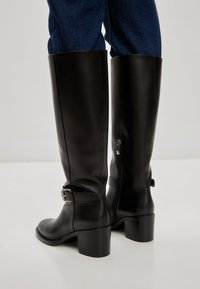 Botine negre din piele, până la genunchi, cu vârf rotund, toc bloc și fermoar lateral. Dotate cu o curea cu cataramă în jurul gleznei. Textură netedă.