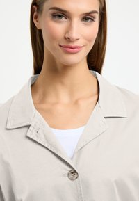 Lichtgrijze jas met een kraag en een enkele knoopsluiting. Zoomachtige stof, eenvoudige ontwerp, gelaagd over een witte top.