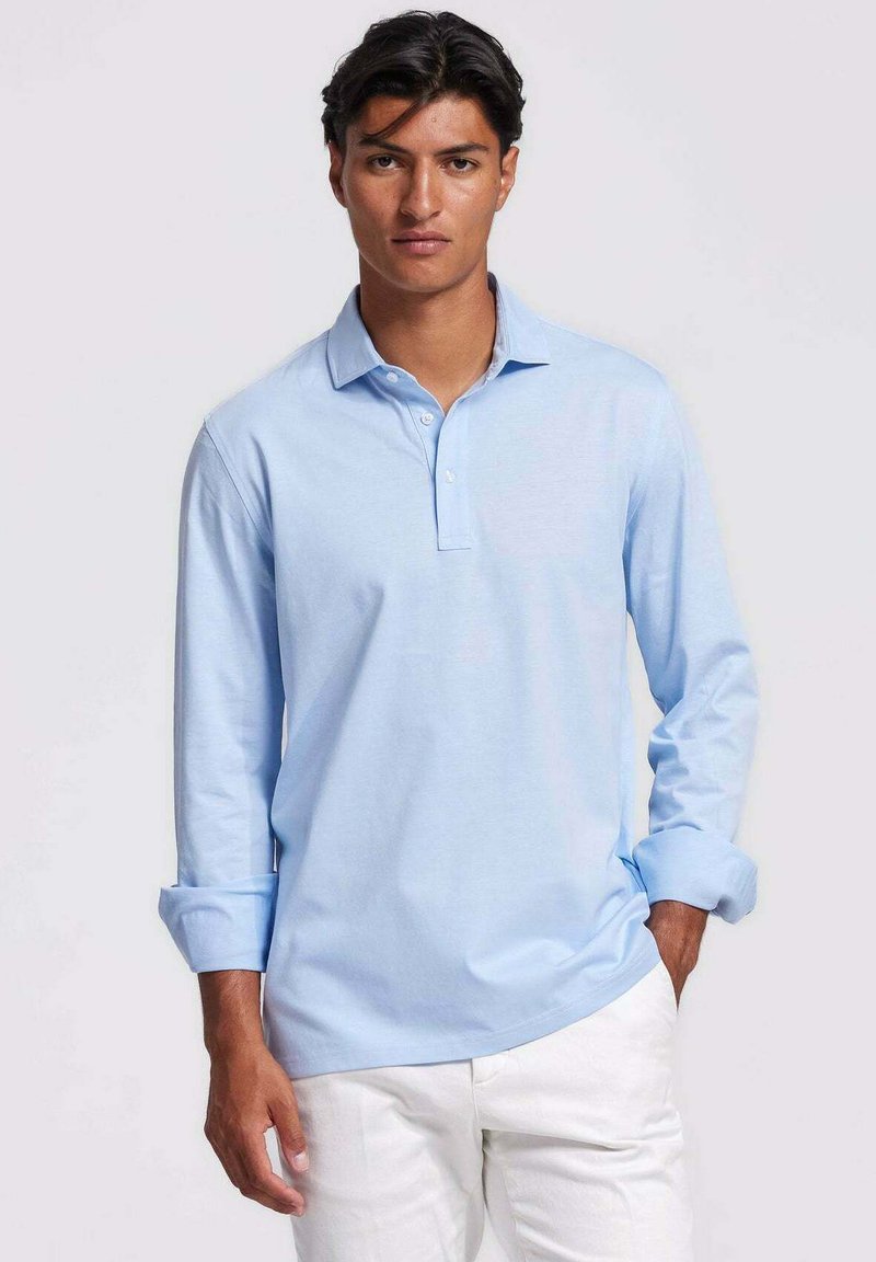 Lichtblauw poloshirt met lange mouwen en een kraag met knopen, gladde textuur en opgerolde manchetten. Draagt bij witte broek.