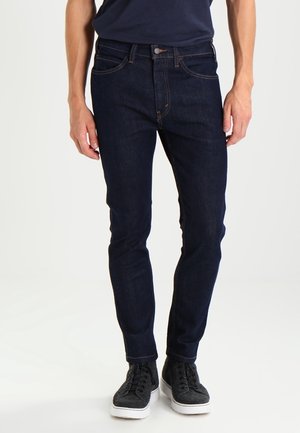 Hombre vistiendo jeans ajustados azul oscuro, camisa azul marino y zapatillas altas gris oscuro, de pie contra un fondo liso.