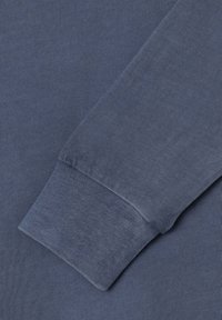 Detailný záber na modrú košeľu s dlhým rukávom, ktorý ukazuje rebrovaný manžetový lem a textúru pleteného materiálu.