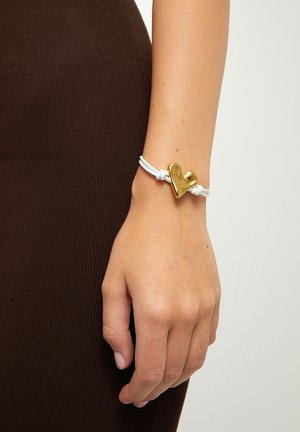Mano de mujer con pulsera de cuerda blanca y charm de corazón dorado irregular sobre fondo de tela marrón.