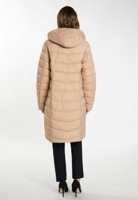 Beige polstret jakke med hætte, med en quiltet struktur, knælængde design og lige pasform. Båret over sorte bukser og hæle.