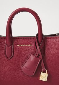 Borsa a tracolla in pelle bordeaux con finitura testurizzata, manici corti e dettagli in logo color oro. Include un piccolo etichetta e un charm con logo.