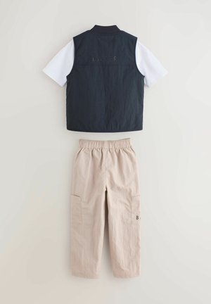 Tumesinine vest valge lühikese varrukaga särgi peal, kombineeritud beežide elastse vöökohaga taskupükstega, laotatud laiale heledale taustale.