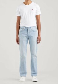 Jeans de denim azul claro con corte recto, combinados con una camiseta blanca básica que presenta un pequeño logo rojo en el pecho. Zapatillas blancas.