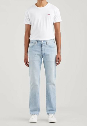 Jean en denim bleu clair avec une coupe droite, associé à un t-shirt blanc uni avec un petit logo rouge sur la poitrine. Baskets blanches.